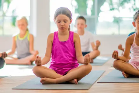 Niños practicando ejercicios de respiración y meditación en un salón de clases, sentados en colchonetas con los ojos cerrados.
