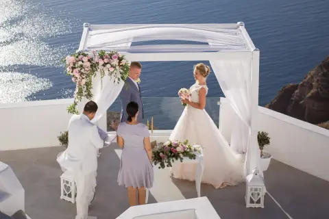 Piękny ślub w Grecji. Para młoda stoi pod białym baldachimem, w tle błękitne morze. Ceremonia odbywa się na tarasie z widokiem na ocean.
