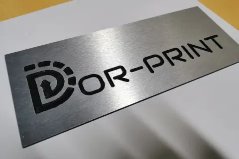 Metalowa tabliczka grawer z logo "Dor-Print". Wygląda jakby była wykonana z aluminium.