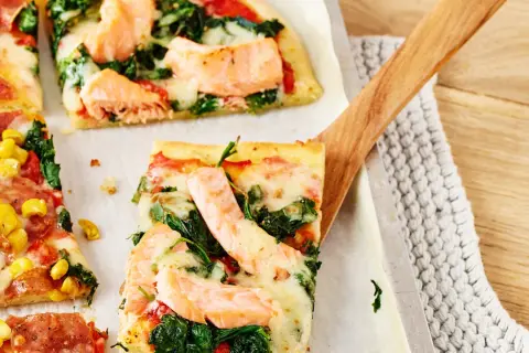 Pizza mit Lachs und Spinat, belegt mit geschmolzenem Käse, wird mit einem Holzschieber serviert.