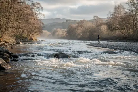 Ein Mann steht am Ufer und geht angeln in Schottland. Der Fluss ist wild und von Bäumen gesäumt.