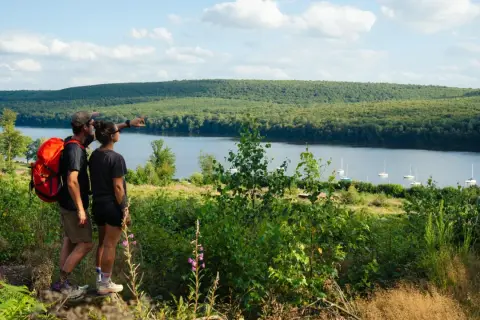 Couple randonneurs admirant le lac des Vieilles Forges, avec des voiliers sur l'eau et une forêt dense en arrière-plan.