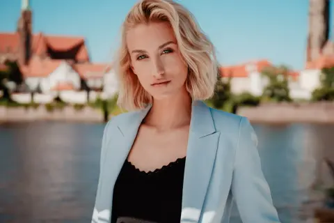 Blondynka w błękitnej marynarce pozuje w plenerze nad rzeką, z widokiem na zabytkowe budynki w tle.