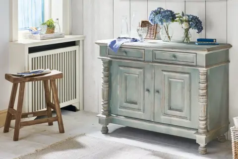 Ein blau-graues Sideboard mit abgenutzter Patina, das den "shabby chic"-Stil verkörpert. Daneben ein Holzhocker und Blumen.