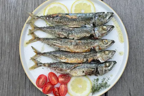 Cinq sardines au four, dorées et savoureuses, servies avec des quartiers de citron, des tomates cerises et des herbes fraîches sur une assiette blanche.