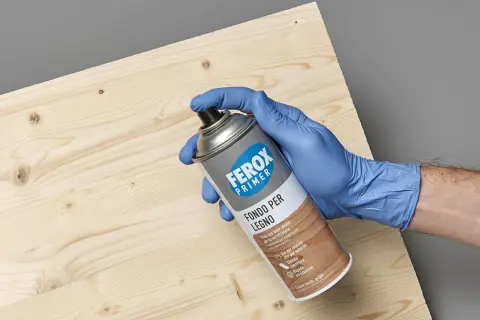 Mano con guanto blu tiene bomboletta spray "Ferox Primer Fondo per Legno", pronta per preparare la superficie.