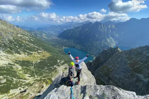 Dwóch wspinaczy na szczycie, z widokiem na Tatry i Morskie Oko. Mnich tatrzański w tle.