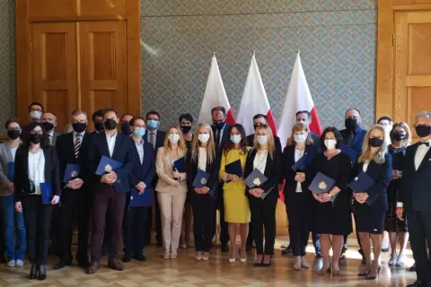 Grupa osób w garniturach i sukienkach, z flagami Polski w tle. Wśród nich radca prokuratorii generalnej, odbierający dokumenty.