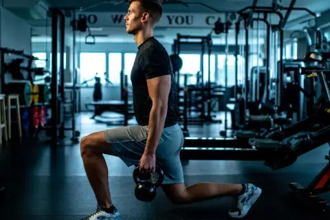 Mann macht Ausfallschritte mit Kettlebell im Fitnessstudio. Verschiedene Ausfallschritte Varianten trainieren Beine und Po.