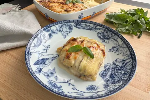 Porzione di lasagne di zucchine gratinate su piatto decorato, con basilico fresco e una teglia sullo sfondo.