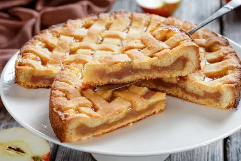 Ein goldbrauner Kuchen mit Gittermuster, gefüllt mit süßem Apfelmus. Ein Stück wird gerade herausgehoben.