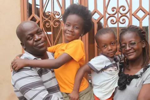 Famille souriante devant une fenêtre ornée. L'homme porte un enfant, la fille étreint son père. L'origine d'Anthony Samama est peut-être liée à cette joie familiale.