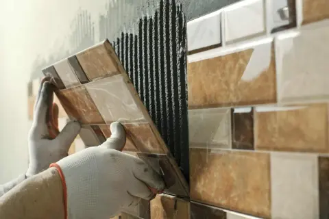 Pose de carrelage sur carrelage, une main gantée appliquant une nouvelle rangée sur le mur déjà carrelé.