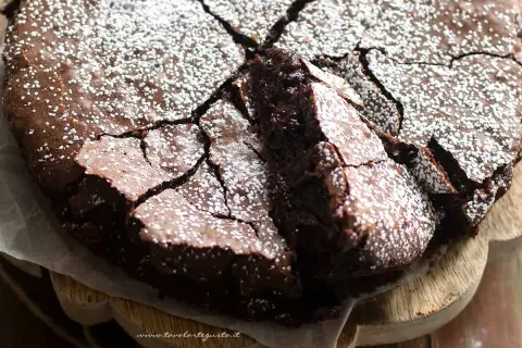 Torta con 2 uova e cioccolato fondente, spolverata di zucchero a velo, con una fetta sollevata che rivela la sua morbidezza.