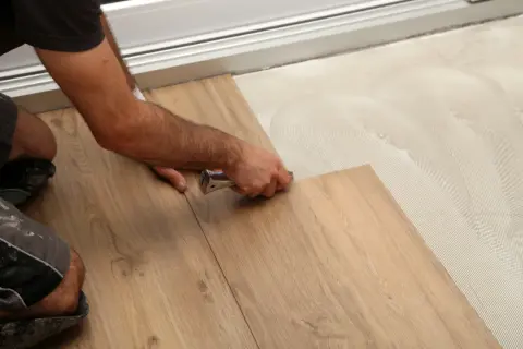 A person installs wood-look drop and lock flooring, using a tool to secure a plank onto adhesive-covered subfloor.