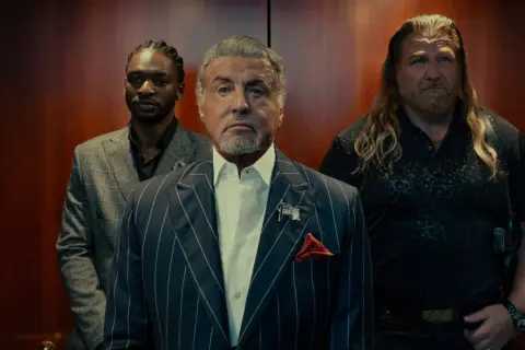 Sylvester Stallone i en serie, flankerad av två män i en hiss.
