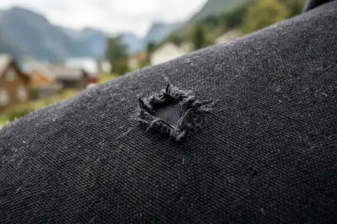 Ett hål i en svart dunjacka, redo för en lagningslapp. I bakgrunden syns norska fjordar och hus.