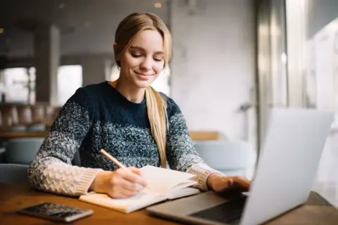 Uśmiechnięta studentka notuje w zeszycie, korzystając z laptopa. Cieszy się z możliwości, jakie dają jej zniżki studenckie.