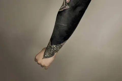 Ein Arm mit einem beeindruckenden **blackout tattoo**, das fast vollständig in tiefem Schwarz gehalten ist. Zarte Mandalas schmücken Handgelenk und Ellenbogen.