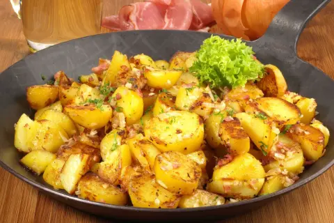 Goldbraune Bratkartoffeln mit Speck und Zwiebeln, garniert mit Petersilie. Ein klassisches Bratkartoffeln Rezept, serviert in einer Eisenpfanne.