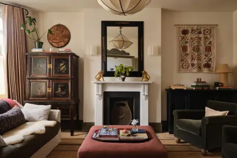 A living room with a plush ottoman, antique cabinet, and fireplace, showcasing a curated collection of minimalist home decor.