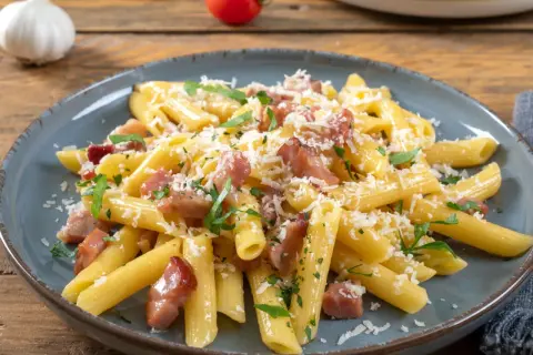 Penne rigate z chrupiącym boczkiem, posypane parmezanem i natką pietruszki. Pyszne makaron z boczkiem na talerzu.
