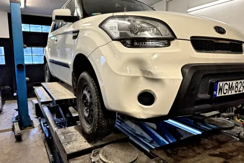 Biały Kia Soul na podnośniku, przygotowany do prac, jak wyciąć otwór na hak holowniczy w dolnej części zderzaka.