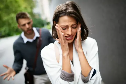 Una donna piange disperata, con le mani sul viso, mentre un uomo alle sue spalle gesticola con frustrazione. L'immagine cattura un momento di profonda infelicità di coppia.