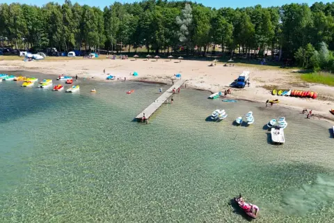 Letnia binduga pole namiotowe nad jeziorem. Woda kusi kajakami, rowerami wodnymi i łódkami. Na plaży odpoczywają ludzie, a w tle widać las.