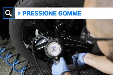 Controllo della pressione gomme auto con manometro. Un tecnico con guanti blu collega lo strumento alla valvola.