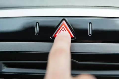 Un doigt appuie sur le bouton des feux de détresse obligatoires, un triangle rouge lumineux sur le tableau de bord d'une voiture.