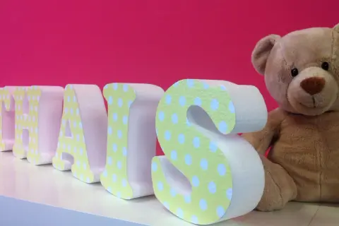 Lettre en relief "THAIS" décorée de pois sur fond rose vif, à côté d'un ours en peluche.