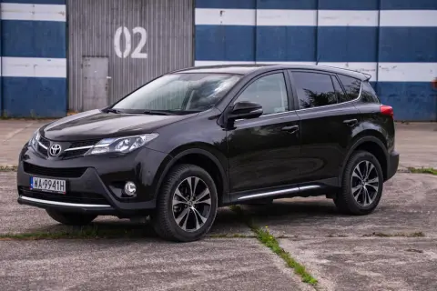 Czarna, używana Toyota RAV4 stoi na betonowym placu przed budynkiem z niebiesko-białymi pasami i numerem "02".