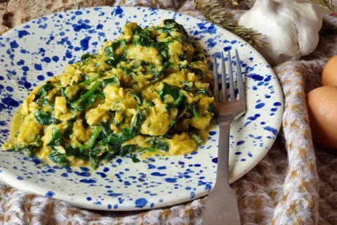 Delicioso revuelto de acelgas con huevo, servido en un plato con salpicaduras azules. Acompañado de pan rústico, ajo y huevos frescos.