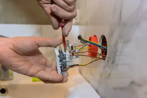 Elektryk montuje gniazdko, łącząc kolorowe kable w puszce. Widać brązowe, żółto-zielone i niebieskie przewody.