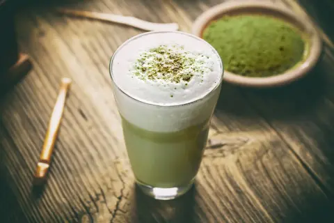 Szklanka z zielonym matcha latte z pianką, obok miseczka z proszkiem matcha i bambusowa trzepaczka.