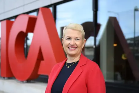 En leende kvinna i röd kavaj framför stora röda bokstäver som bildar ICA. Hon är ICA:s VD.