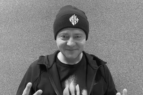 Mężczyzna w czapce z logo, w bluzie i koszulce. W tle szara ściana. Nie wiadomo, **joka na co zmarł**.
