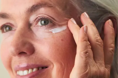 Eine Frau um die 60 trägt Augencreme auf. Ihre Haut zeigt natürliche Spuren des Alters, aber keine Pickel.