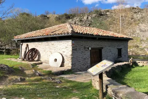 El molino de la Hiruela, con su tejado de tejas rojas y muros de piedra, se alza junto a un río. A su lado, piedras de moler y una rueda de hierro evocan su pasado.