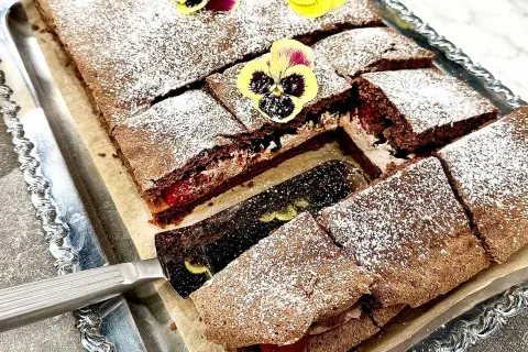 En chokladgrädde tårta skuren i bitar, pudrad med florsocker och dekorerad med violer. En tårtspade lyfter upp en bit.
