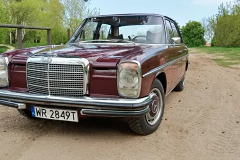 Klasyczny mercedes w114 w bordowym kolorze, zaparkowany na piaszczystej drodze wśród zieleni.
