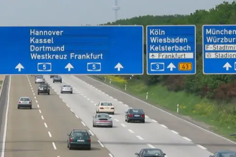 Niemiecki **autobahn** z niebieskimi tablicami kierunkowymi do Hanoweru, Kolonii i Monachium. Samochody jadą po wielopasmowej drodze.