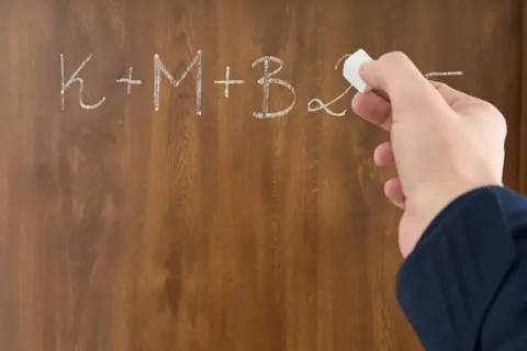 Dłoń pisze kredą na tablicy: K+M+B. Nagle przychodzi epifania, rozwiązanie problemu.