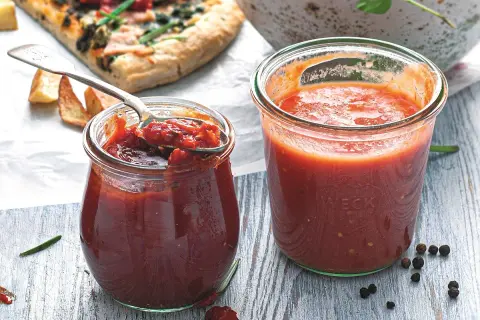 Dwa słoiki domowego sosu do pizzy, jeden z łyżką pełną aromatycznej masy. Obok kawałek pizzy i przyprawy.