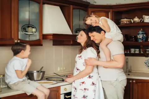 Rodzina w kuchni przygotowuje naleśniki. Tata niesie córkę na barana, mama miesza ciasto, a synek siedzi na blacie. LZO co to? To rodzinne chwile!