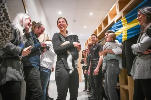Elin Johansson, med ett leende och en bebis i bärsele, omgiven av glada människor och en svensk flagga. Kanske en paus från taekwondo-träningen?