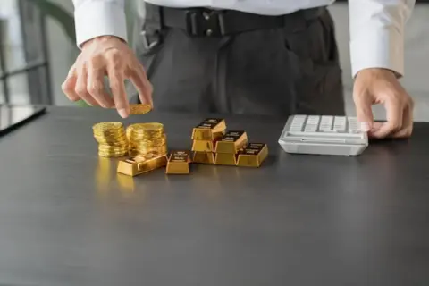 Ręce układają stosy złotych monet i sztabek, obok kalkulator. To inwestycja w złoto Heraeus.