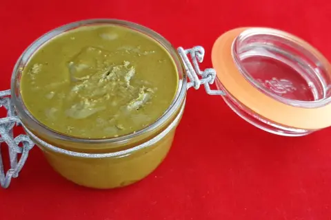 Un pot en verre ouvert révèle une délicieuse pâte à tartiner pistache maison, d'une belle couleur verte, sur un fond rouge vif.