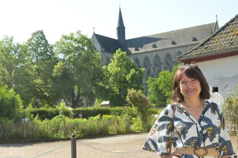 Frau lächelt vor einem Dom, umgeben von Bäumen und einem Garten. Ein Ort, der zum Verweilen und zur Entdeckung von Literatur am Dom einlädt.
